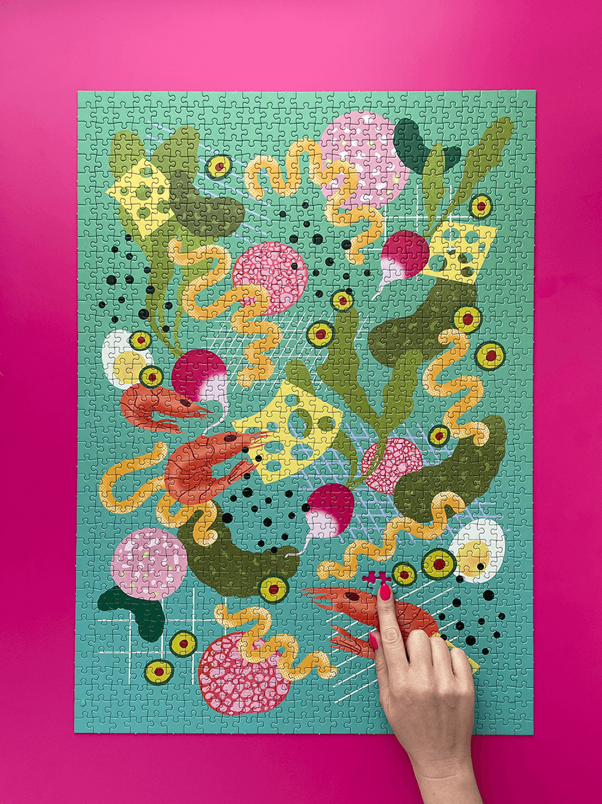 Ein fertiges "Picky Bits" Puzzle liegt auf pinkem Untergrund, eine Hand mit pinken Nägeln kommt von unten ins Bild und legt das letzte Teil.